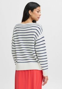 Weißer Strickpullover mit navyblauen horizontalen Streifen, ausgestattet mit gerippten Bündchen und Saum. Kombiniert mit einem fließenden roten Rock.