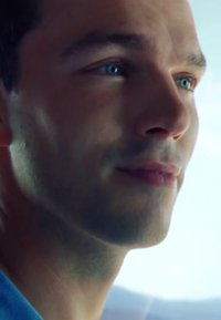 Close-up of a male face with smooth skin, light stubble, and bright blue eyes. He wears a blue shirt, with a neutral expression.