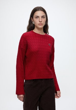 Jeune femme aux longs cheveux bruns portant un pull rouge en maille torsadée avec un petit détail en forme de cœur et un pantalon foncé, debout devant un fond uni.
