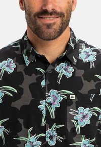 Cămașă cu mâneci scurte dintr-un material negru, cu un model floral care include flori albastre și frunze verzi. Design cu nasturi și un buzunar frontal.
