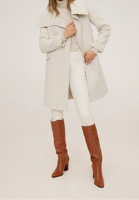 Abrigo gris claro con gran solapa, mangas largas y bolsillos laterales, combinado con jeans blancos y botas de cuero marrón hasta la rodilla.