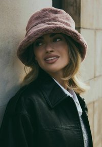 Chapeau seau en peluche rose, texture douce, forme ronde. Porté avec une veste en cuir noire et une chemise blanche à col, sur un fond neutre.