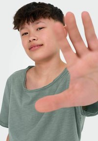 Jasnozielona koszulka z krótkim rękawem o okrągłym dekolcie i surowym wykończeniu, noszona przez dziecko wyciągające rękę w stronę widza.