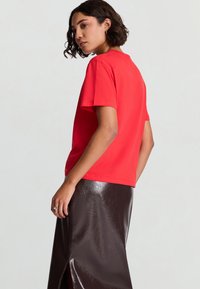 Maglietta rossa di cotone con maniche corte, abbinata a una gonna marrone lucida dalla trama lavorata con spacco laterale. Design semplice, vestibilità comoda.