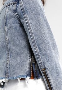 Jeansjacka med fransad nederkant och dragkedjedetalj på ärmen, bärs över vita byxor och visar sidoprofil och bakvy.