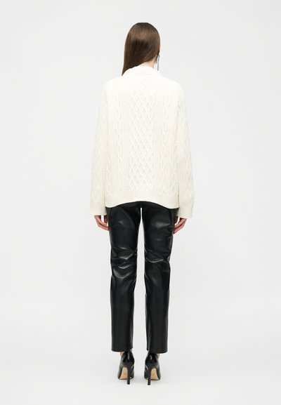Γυναίκα με την πλάτη γυρισμένη, που φοράει λευκό πουλόβερ πλεκτό με πλεξούδες, μαύρο δερμάτινο παντελόνι και μαύρες γόβες, μπροστά σε ένα απλό φόντο.