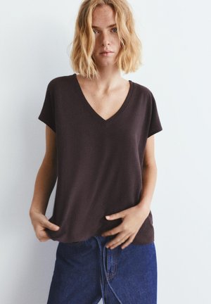 Junge Frau mit kurzen blonden Haaren trägt ein schwarzes T-Shirt mit V-Ausschnitt und einen blauen Jeansrock, steht vor einem schlichten weißen Hintergrund.