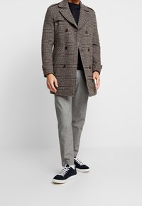 Manteau croisé en laine à carreaux marron avec larges revers, boutons marron, porté sur une chemise noire et un pantalon gris à motifs, associé à des baskets bleu marine.