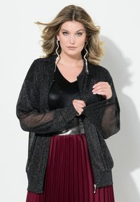 Giacca bomber nera con texture scintillante, maniche trasparenti, polsini a costine e chiusura con zip; abbinata a una gonna plissettata color bordeaux intenso.