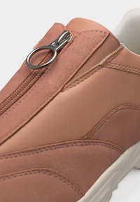 Gros plan sur une basket beige avec des panneaux en suède et en cuir, équipée d'une fermeture éclair centrale avec une tirette métallique ronde et une semelle blanche.