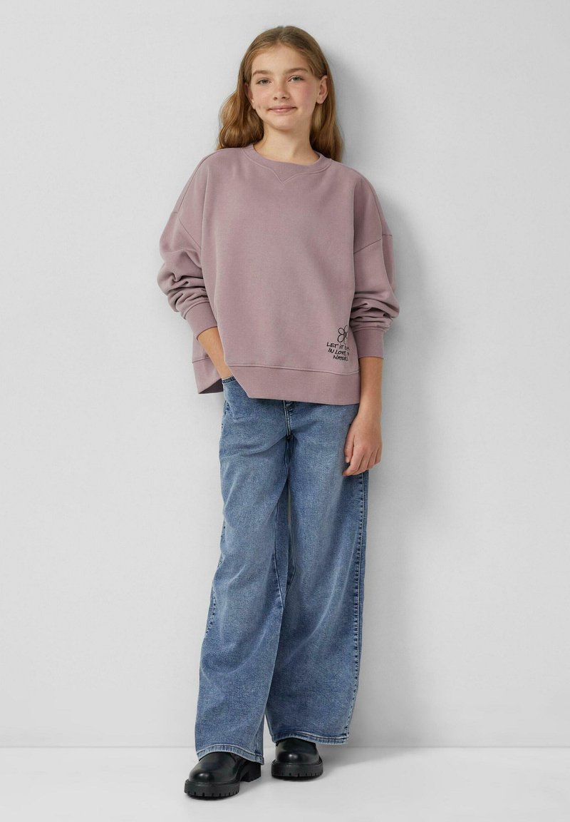 Różowy, oversize'owy sweter z okrągłym dekoltem, z małym kwiatowym nadrukiem na dolnym brzegu. Połączony z niebieskimi spodniami o szerokich nogawkach i czarnymi klapkami.