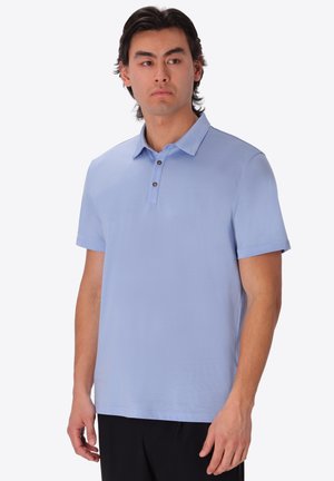 Giovane uomo con capelli scuri di media lunghezza, che indossa una polo azzurra a maniche corte e pantaloni neri, che guarda leggermente di lato.
