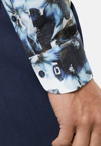 Shirt mouw met een bloemenpatroon in blauw en zwart, op een witte achtergrond, enkele knoopsluiting en een gestructureerde donkerblauwe stof.