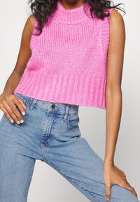 Top sin mangas de punto rosa con cuello alto y dobladillo acanalado, combinado con jeans de mezclilla azul claro. Manos en las caderas.