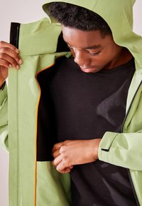 Hellgrüne wasserfeste Jacke mit Kapuze und orangenem Saum. Das Innere ist schwarz, mit einer sichtbaren Tasche und glatter Textur.