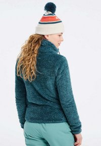 Mėlynos spalvos fliso striukė su aukštu apykakle, ilgomis rankovėmis ir tekstūruota apdaila; derinama su balta megzta kepure, turinčia mėlynas ir oranžines juostas.