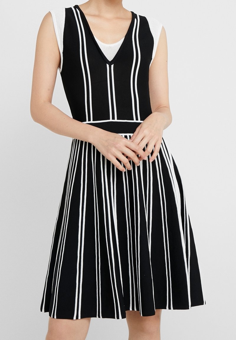 Femme portant une robe noire sans manches avec des rayures verticales blanches et un col en V sur un haut en résille blanc, les mains doucement jointes devant.