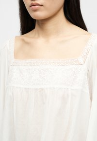 Femme portant un chemisier blanc avec un col carré, une bordure en dentelle et une broderie florale sur la poitrine.