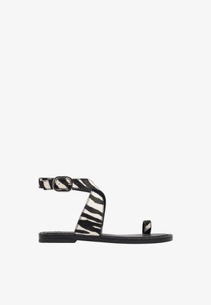 Płaska sandałka z czarno-białymi paskami w zebra print, klamrą przy kostce i pętlą na palec na czarnej podeszwie.