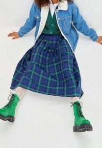 Chaqueta de mezclilla con cuello de peluche sintético, chaleco de punto verde, falda a cuadros en azul y verde, botas verdes con suelas gruesas y calcetas, calcetas de red.