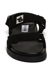 Svart sandal med två velcro-remmar, en vadderad fotbädd och en elegant silverfärgad spänne. Tillverkad av mjuka, strukturerade material.