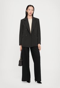 Blazer doppiopetto grigio scuro con ampie rever, indossato su un top beige, abbinato a pantaloni larghi neri e tacchi a punta neri.