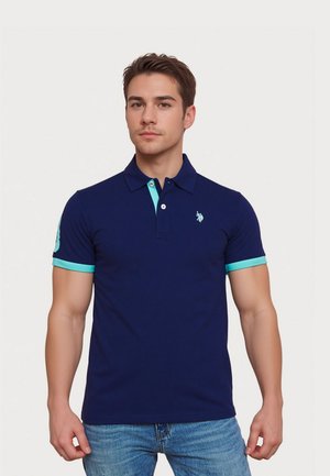 Jeune homme debout, portant un polo bleu marine avec des bordures turquoise et un jean bleu clair sur un fond blanc uni.