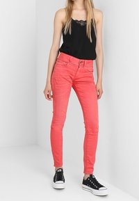 Jean skinny rosa de denim, con cinco bolsillos y pasadores para cinturón, combinados con una camiseta sin mangas con encaje negro y zapatillas negras.