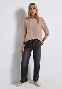 Lichtroze blouse met lange mouwen en een ronde halslijn, gecombineerd met loszittende donkergrijze jeans en beige sneakers met accenten.