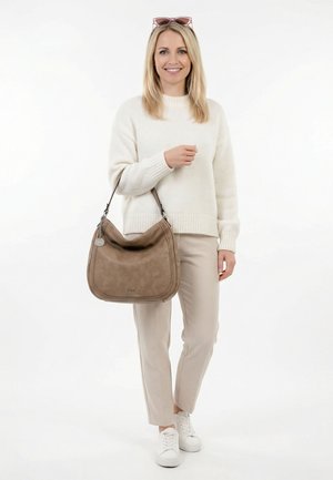Donna sorridente con maglione crema e pantaloni beige tiene una grande borsa marrone, indossa scarpe da ginnastica bianche e occhiali da sole sulla testa.