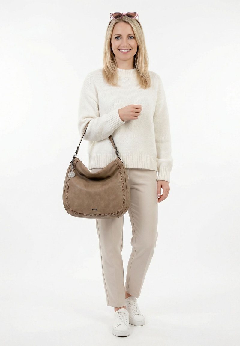 Smiling woman in cream sweater and beige pants holds a large brown handbag, wearing white sneakers and sunglasses on her head.