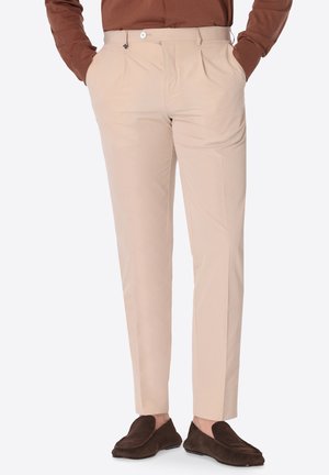 Pantaloni beige su misura indossati con mocassini in suede marroni e camicia a maniche lunghe color ruggine, mani in tasca, in piedi su sfondo semplice.