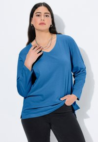 Blaues, langärmliges Oberteil mit V-Ausschnitt, aus weichem Material gefertigt. Verfügt über eine lockere Passform, seitliche Schlitze und eine geschichtete, silberfarbene Halskette.