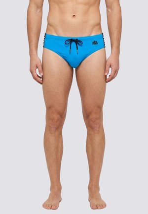 Uomo che indossa slip da nuoto blu aderenti con coulisse nera e logo sul davanti, in piedi scalzo su uno sfondo chiaro e uniforme.