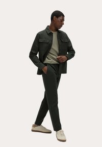 Uomo con giacca e pantaloni verde scuro, camicia verde chiaro e sneakers beige, in piedi con una mano in tasca e lo sguardo rivolto verso il basso.