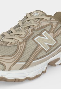 Μπεζ και καφέ αθλητικό παπούτσι New Balance με υφασμάτινη υφή, πλαστικές επικαλύψεις και λευκή σόλα, που δείχνει λεπτομέρειες από μπροστά και πλευρά.