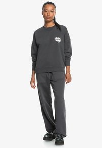 Mørkegrå sweatshirts med lange ermer, rund hals og en liten logo på brystet, parret med matchende bukser og svarte sko.
