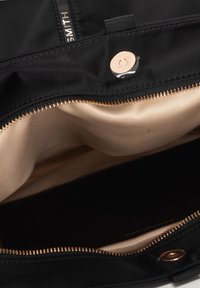Interno di una borsa nera in nylon con fodera in satin oro. Presenta una chiusura a zip con accessori in oro rosa e un dettaglio con bottone a pressione magnetico.