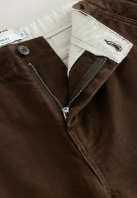 Brown relaxed-fit pants with partially open zipper, button closure, and white inner waistband with size label.