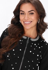 Chaqueta negra con cremallera, con tejido texturizado, adornos de perlas y piedras brillantes en el cuello y por toda la prenda, y un diseño estructurado.