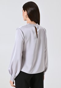 Blusa in raso grigio chiaro con chiusura a forma di occhiello sul retro, maniche lunghe a palloncino e un leggero orlo asimmetrico, abbinata a pantaloni neri.