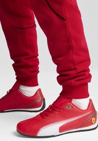 Czerwone sportowe buty z białymi akcentami, gładka skóra, czarna podeszwa i logo Ferrari po boku, w zestawie z czerwonymi spodniami dresowymi z mankietami.