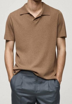 Mann trägt ein braunes, kurzärmeliges Strick-Poloshirt, in eine hochgeschnittene graue plissierte Hose gesteckt, mit Händen in den Taschen.