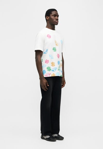 Άνδρας που στέκεται φορώντας λευκό μπλουζάκι με πολύχρωμα τυπώματα από σύμβολα δολαρίου και διαμάντια, μαύρο παντελόνι και μαύρα αθλητικά παπούτσια με λευκές ρίγες.