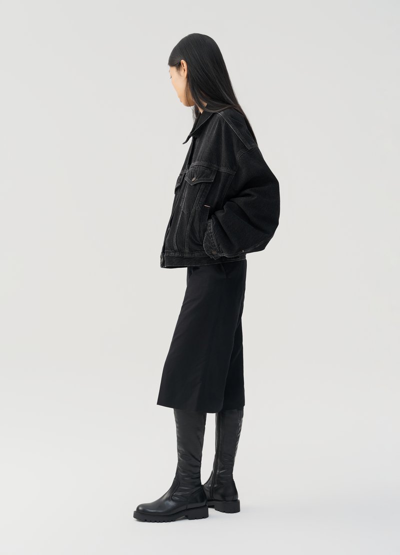 Giacca di jeans nera con una vestibilità rilassata, abbinata a una gonna midi nera e stivali neri alti fino al ginocchio. Il modello è in piedi di lato su uno sfondo neutro.