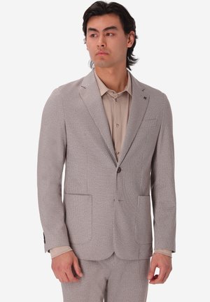 Uomo che indossa un abito grigio chiaro strutturato con pantaloni abbinati e camicia beige con bottoni, che guarda leggermente di lato contro uno sfondo bianco.