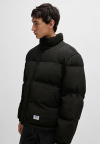 Schwarze Daunenjacke mit hohem Kragen, großen quilted Abschnitten und einem vorne befindlichen Reißverschluss. Verfügt über eine linke Tasche mit einem Logoschild.