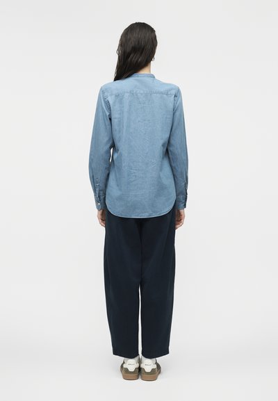 Άτομο με μακριά σκούρα μαλλιά που φοράει ανοιχτό μπλε πουκάμισο από τζιν, σκούρο παντελόνι και λευκά αθλητικά παπούτσια, στεκόμενο με την πλάτη προς την κάμερα σε απλό φόντο.