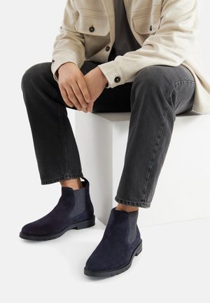 Granatowe zamszowe botki z elastycznymi panelami po bokach i teksturowanym wykończeniem, w połączeniu z ciemnymi dżinsami i jasną kurtką.