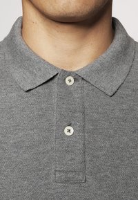 Harmaa polo-paita, jossa on kauluskotelo, sisältää kaksi nappia ja on valmistettu teksturoidusta kankaasta, keskittyen kaulus- ja nappitietoihin.